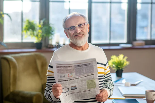 Ősz hajú, szakállas férfi, aki szépen mosolyog, miközben újságot olvas. — Stock Fotó