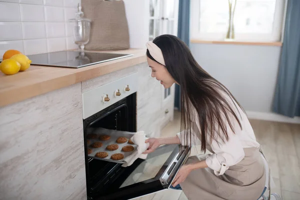 Chica joven ama de casa sentada cerca del horno sacando bandeja para hornear . — Foto de Stock