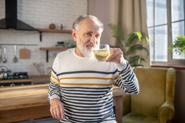 Sakallı, gri saçlı, çizgili kazaklı, çay içen adam.