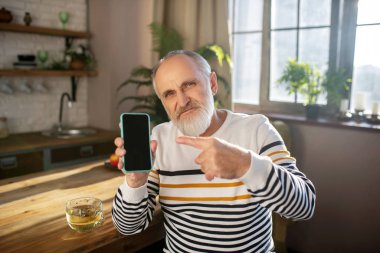 Sakallı, gri saçlı, çizgili kazaklı adam akıllı telefonunu gösteriyor.