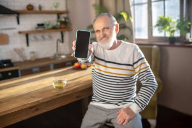 Sakallı, gri saçlı, çizgili kazaklı adam mutfakta oturuyor.