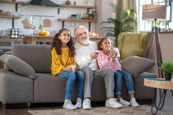 Deux filles à la peau foncée et leur grand-père regardant la télévision ensemble assis sur le canapé — Photo