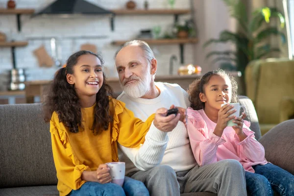 Två mörkhyade flickor och deras farfar tittar på tv tillsammans — Stockfoto