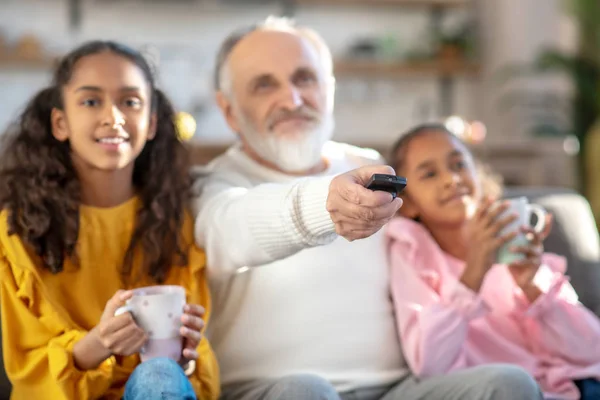 Dos chicas de piel oscura viendo televisión con su abuelo y sonriendo —  Fotos de Stock