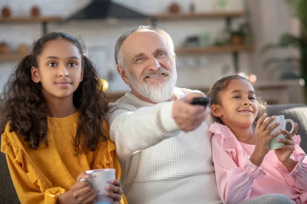 Dos chicas de piel oscura sentadas junto a su abuelo y viendo la televisión —  Fotos de Stock