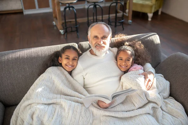 Abuelo y sus dos nietas sentados en el sofá y sintiéndose cómodos —  Fotos de Stock