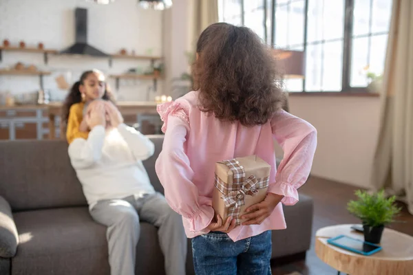 Menina de pele escura em uma camisa rosa escondendo um presente de aniversário para seu avô nas costas — Fotografia de Stock