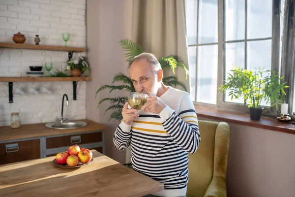 Vousatý šedovlasý muž v pruhovaném svetru, který si dává doma čaj — Stock fotografie