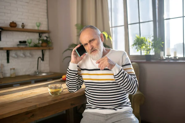 Sakallı, gri saçlı, çizgili kazaklı bir adam mutfakta oturuyor ve telefonda konuşuyor. — Stok fotoğraf