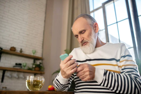 Hombre de pelo gris barbudo en un suéter a rayas sentado en la cocina y mirando su teléfono — Foto de Stock