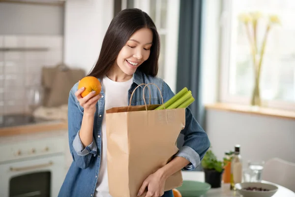 Vacker kvinna med orange i handen tittar in i väskan med grönsaker. — Stockfoto