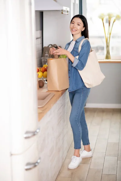 Νεαρή γυναίκα στέκεται με σακούλες στην κουζίνα. — Φωτογραφία Αρχείου