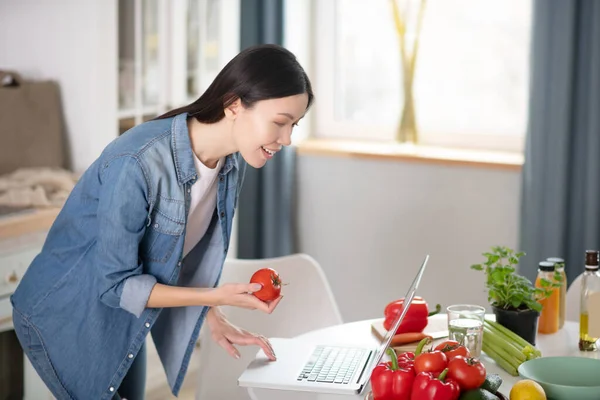 Elegante chica joven de pie cerca de la computadora portátil con tomate en la mano . — Foto de Stock