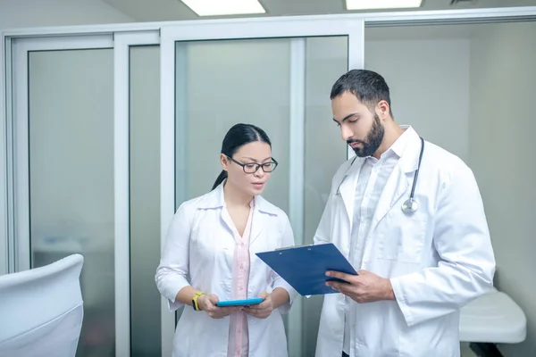 Dark-haired male and female doctors standing in the room together discussing schedule — Stock Fotó