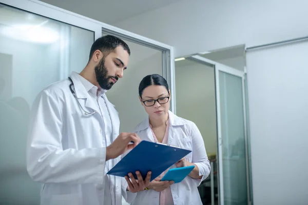 Dark-haired male and female doctors standing in the room together and looking busy — Stock fotografie