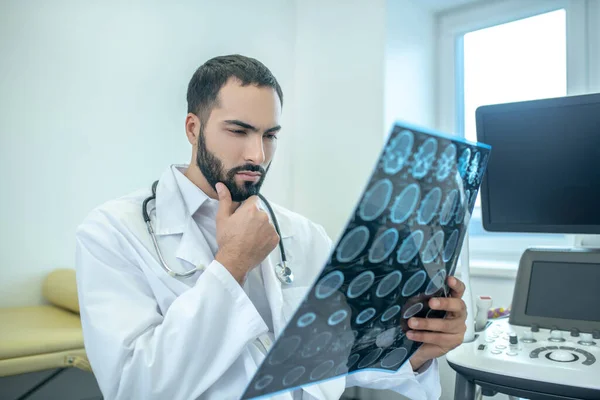 Médico barbudo masculino con una bata blanca que analiza los resultados de la resonancia magnética con aspecto grave — Foto de Stock
