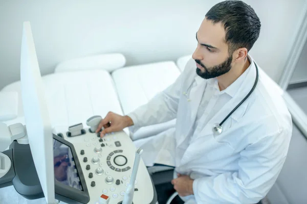 Médico barbudo masculino con una bata blanca sentado en el dispositivo ultrasónico que parece ocupado —  Fotos de Stock