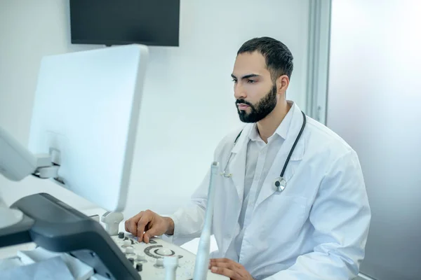 Maschio medico barbuto in vestaglia bianca seduto al dispositivo a ultrasuoni — Foto Stock