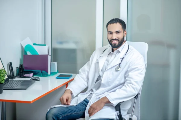 Médico varón con una bata blanca sentado en su consultorio — Foto de Stock