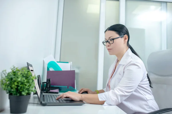 Female dark-haired doctor in eyewear working on a laptop — Φωτογραφία Αρχείου