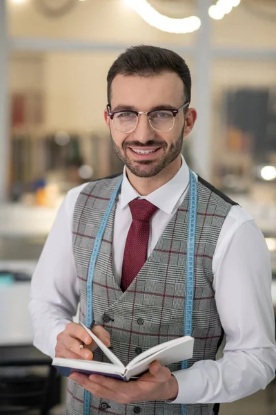 Male tailor wearing glasses, holding notebook, smiling — ストック写真