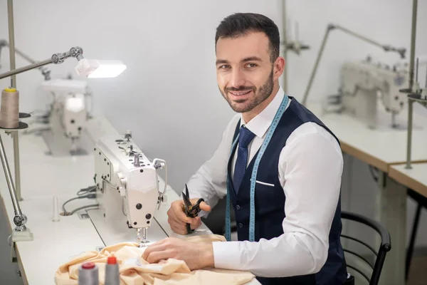 Male tailor sitting at sewing machine, holding scissors — ストック写真