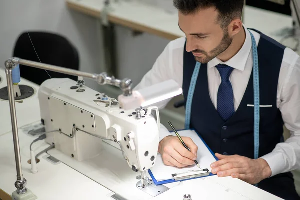 Alfaiate masculino sentado na máquina de costura, fazendo anotações — Fotografia de Stock