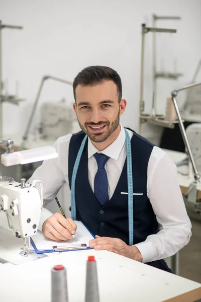Male tailor sitting at sewing machine, making notes, smiling — Stockfoto