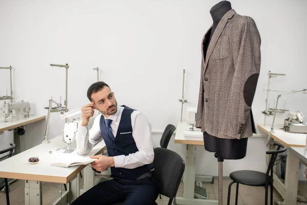 Male tailor sitting with book, looking at jacket — ストック写真