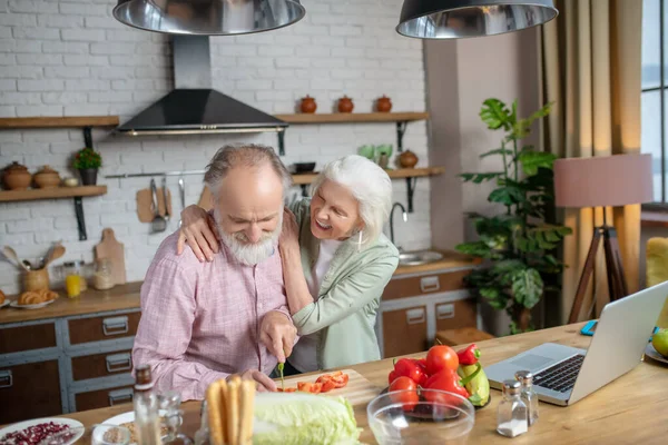 Ältere Frau umarmt ihren Mann, während er Tomaten schneidet — Stockfoto