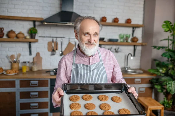 Smiling pensioner wearing apron holding just baked cookies — Stock Photo, Image