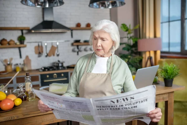Mujer mayor leyendo un periódico en su cocina — Foto de Stock