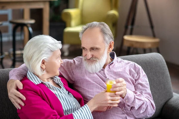 Grey-haired bearded man giving a glass of orange juice to his wife and smiling — Stock Photo, Image