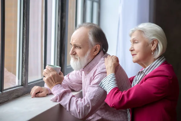 Elderly grey-haired couple standing near the window looking thoughtful — Zdjęcie stockowe