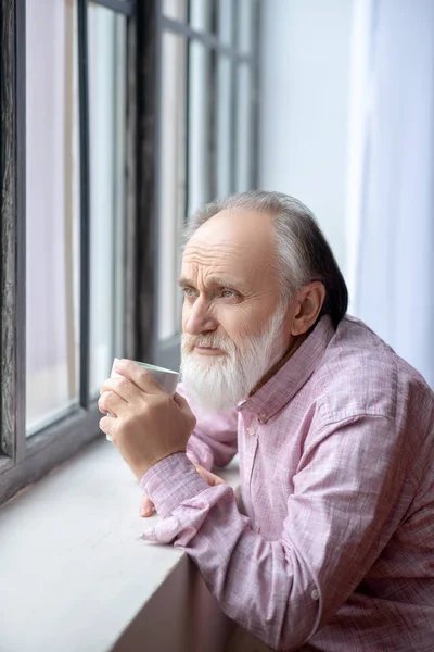 Cabelo grisalho barbudo em pé perto da janela olhando pensativo — Fotografia de Stock