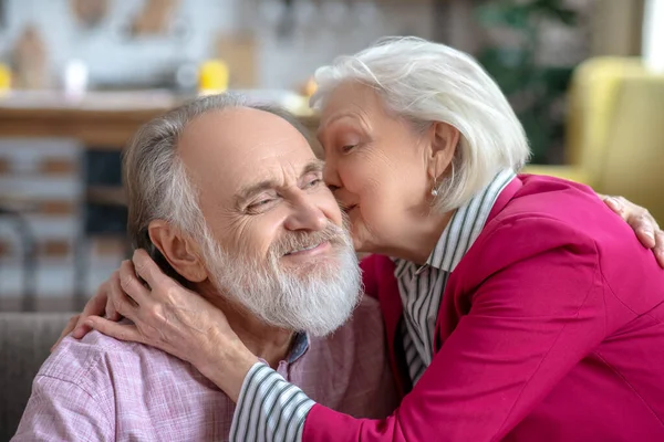 Elderly greay-haired woman kissing her happy bearded husband on a cheek — 스톡 사진