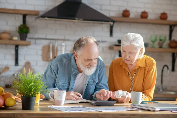 Elderly married couple sitting in the kitchen and making calculations — 图库照片