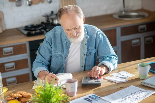 Homem de cabelos grisalhos barbudo sentado à mesa e fazendo cálculos — Fotografia de Stock