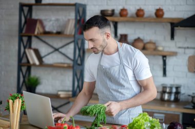 Man in an apron with greens in hand looking at laptop.