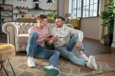Two men with cups sitting side by side on the floor.
