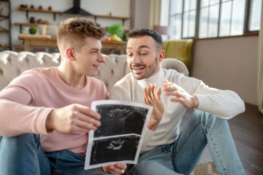Male couple with pictures of ultrasound sitting on floor at home.