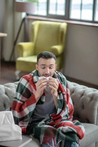 Dark-haired young sick man covered with plaid sneezing — Stockfoto