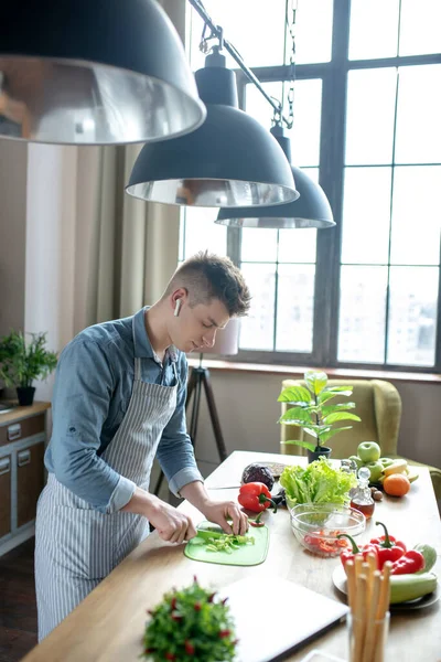 Blond-haired man in wireless headphones and apron preparing salad. — Stockfoto