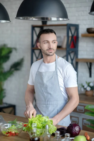 Dark-haired man in a kitchen apron cooking at home. — Stockfoto