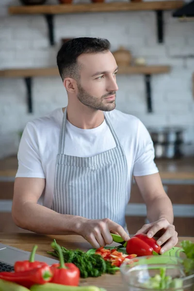 Good looking young man with a beard slicing pepper at home. — Stock fotografie
