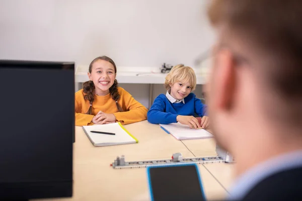 Учні сидять за столом навпроти вчителя, посміхаючись — стокове фото