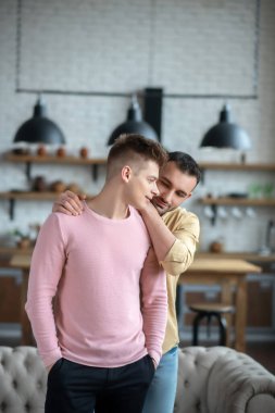 Koyu renk saçlı, turuncu gömlekli bir adam ortağına sarılıyor.