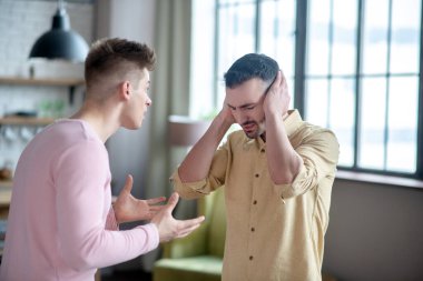 Ortağı ona bağırırken turuncu gömlekli adam kulaklarını kapatıyor.