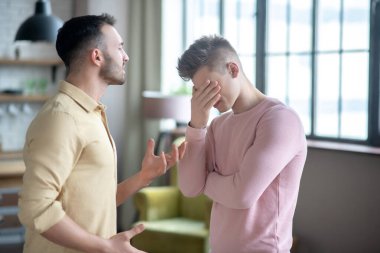 Koyu saçlı, turuncu gömlekli bir adam ortağını suçluyor.