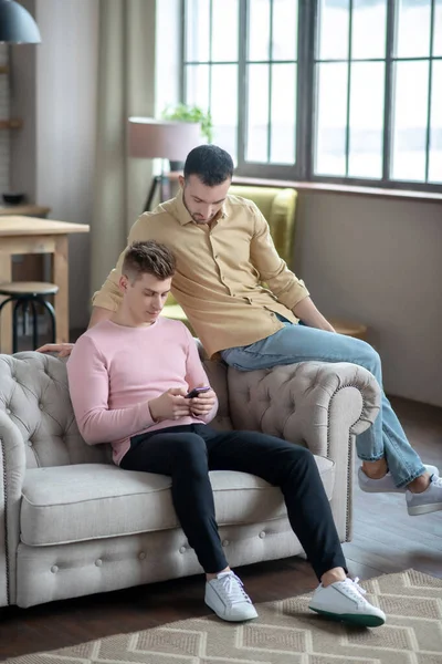 Man in orange shirt sitting on the sofa watching his partner chatting online — Stock Photo, Image
